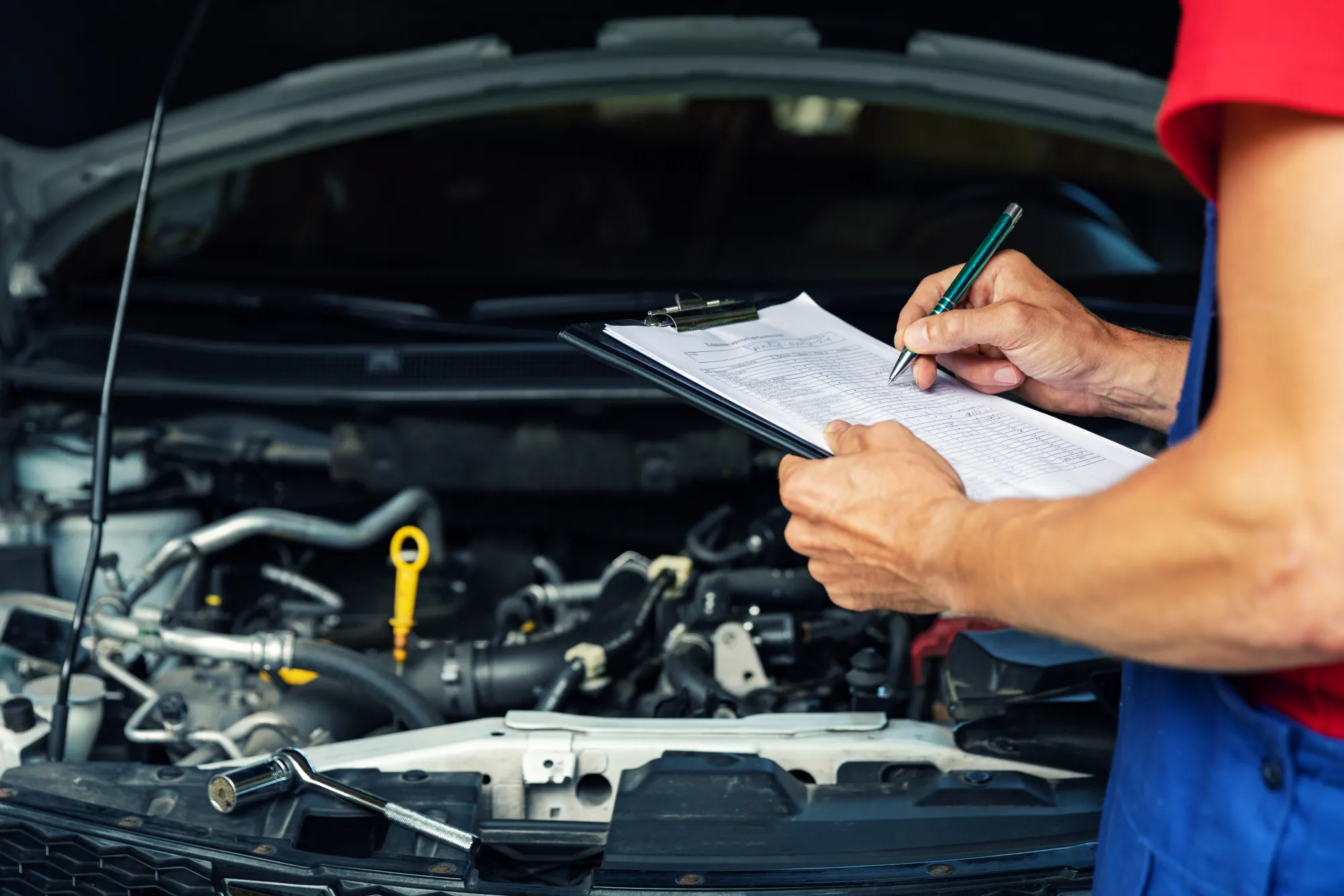A mechanic in a red shirt and blue overalls writes on an inspection checklist clipboard in front of a car's open hood and engine bay.